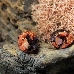 May include: A pair of round, resin earrings with a mix of pink, orange, and purple colors. The earrings are embedded with copper flakes and have a shiny, translucent finish.