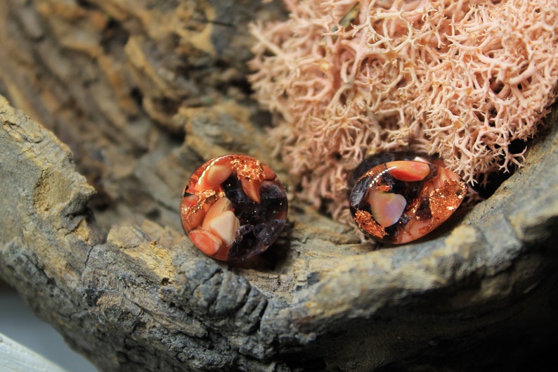 May include: A pair of round, resin earrings with a mix of pink, orange, and purple colors. The earrings are embedded with copper flakes and have a shiny, translucent finish.