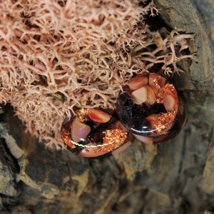 May include: Two round, clear glass plugs with copper flakes and pink and white resin pieces. The plugs are on a brown, textured surface.