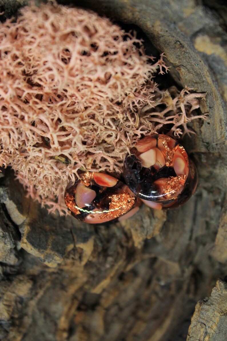 May include: Two round, clear glass plugs with copper flakes and pink and white resin pieces. The plugs are on a brown, textured surface.