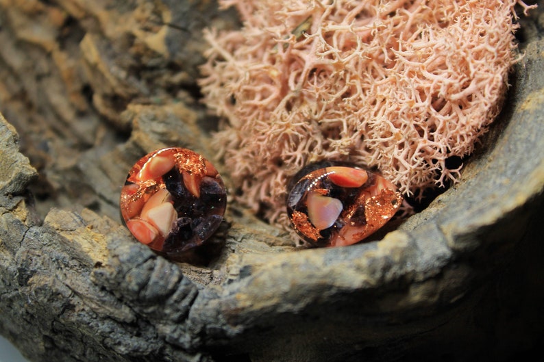 May include: Two round resin earrings with a mix of copper, pink, and black accents. The earrings are displayed on a piece of wood.