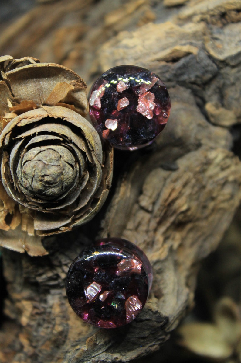 May include: Two round glass plugs with a dark purple base and embedded pink and gold glitter.