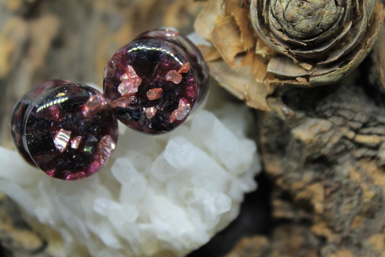 May include: Two round, dark purple glass plugs with embedded iridescent pink and gold glitter.