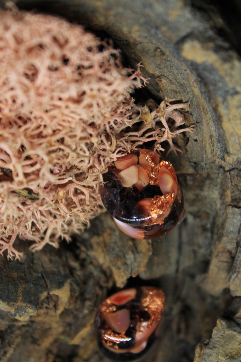 May include: Two round, clear glass ornaments with copper foil and pink and white pieces inside. The ornaments are sitting on a piece of tree bark.