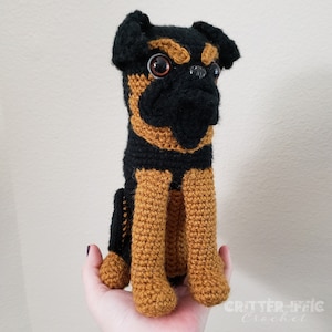 May include: Crocheted stuffed toy dog with black and brown fur. The dog is sitting with its legs crossed and has large brown eyes.