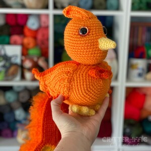 May include: A crocheted orange bird with yellow feet and a yellow beak. The bird has large black eyes and a fluffy orange tail.