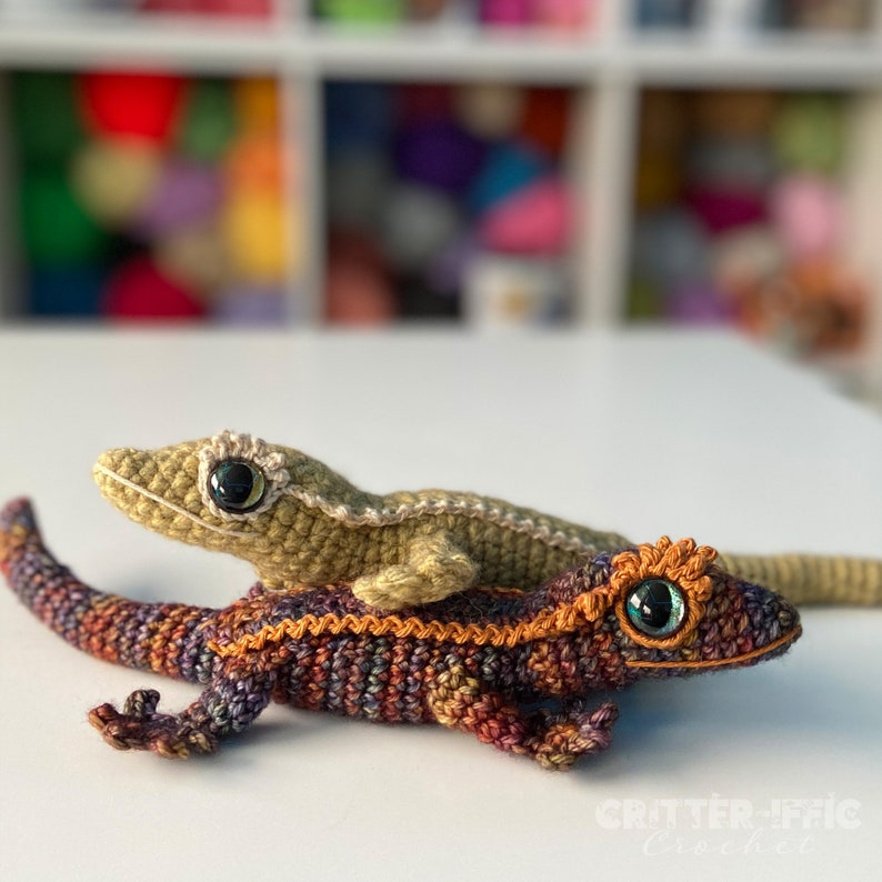 two crocheted geckos sitting on a table , one brown and orange and one gold with wide eyelashed eyes