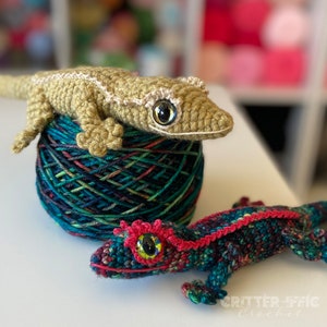 Two crocheted crested geckos one gold and one teal with red accents sitting on a table