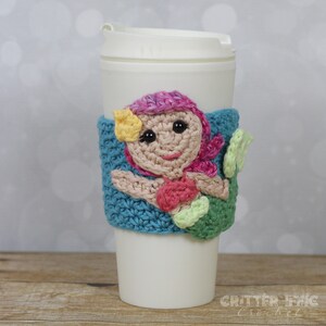 May include: A crocheted mermaid coffee cup cozy with a pink and yellow mermaid with black eyes and a green tail. The cozy is turquoise blue.