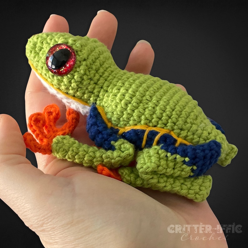 crocheted red eyed tree frog sitting on a hand looking left on a black background