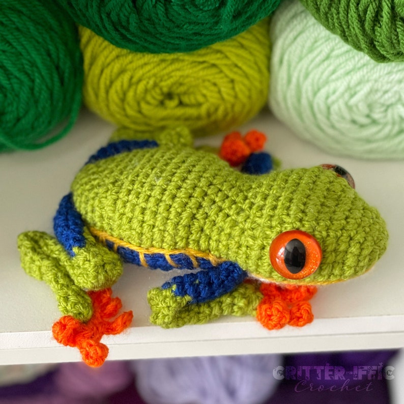 May include: A green crocheted frog with blue and yellow accents, orange feet, and red eyes. The frog is sitting on a white surface.