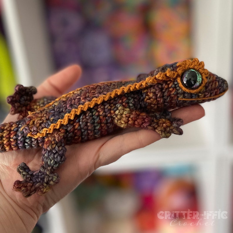 crocheted crested gecko being held by a hand in front of a blurred background. Brown and orange with bright eyes