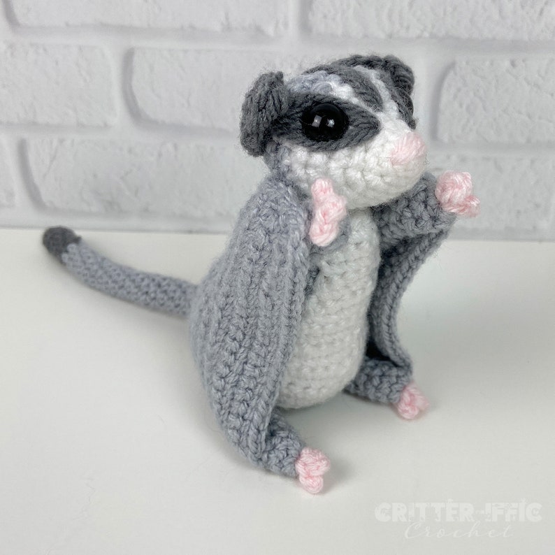 grey crocheted sugar glider sitting on a white table near a white brick wall