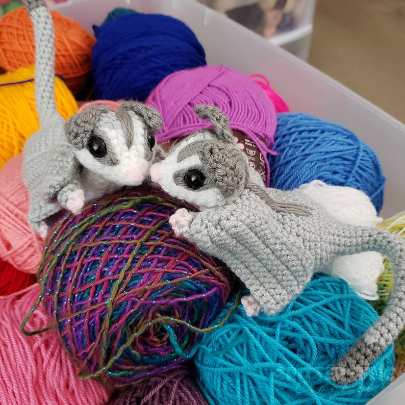 two grey crocheted sugar gliders sitting in a bin full of yarn playfully