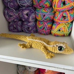 a gold crocheted crested gecko sitting on a white shelf in front of colorful yarn