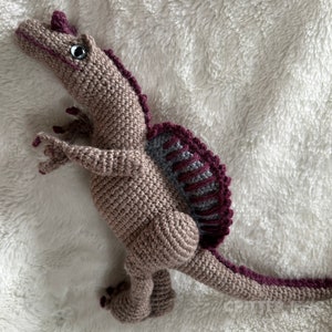 Pode incluir: Um brinquedo de dinossauro Spinosaurus de croch&ecirc; feito &agrave; m&atilde;o. O brinquedo &eacute; em tons de bege, cinza e bord&ocirc;. Possui um corpo texturizado, uma vela distinta e membros pequenos e detalhados. O dinossauro tem uma cauda longa.