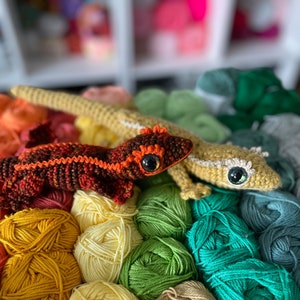 two crocheted crested geckos, one orange and one gold sitting on a colorful yarn background