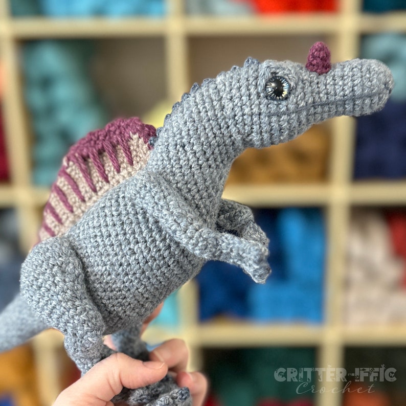 Pode incluir: Um brinquedo de dinossauro de croch&ecirc; em tons de cinza, com uma vela roxa e bege nas costas e um pequeno chifre roxo. O dinossauro tem olhos grandes e detalhados e &eacute; segurado na m&atilde;o de uma pessoa. O texto "Critter-iffic Crochet" &eacute; vis&iacute;vel.