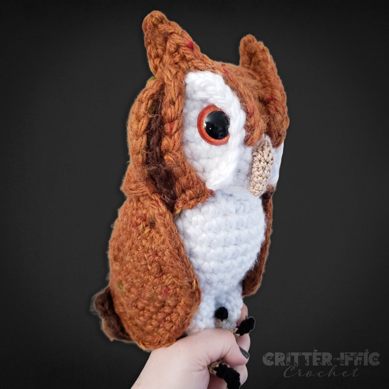 realistic crocheted screech owl being held by a hand in front of a black background