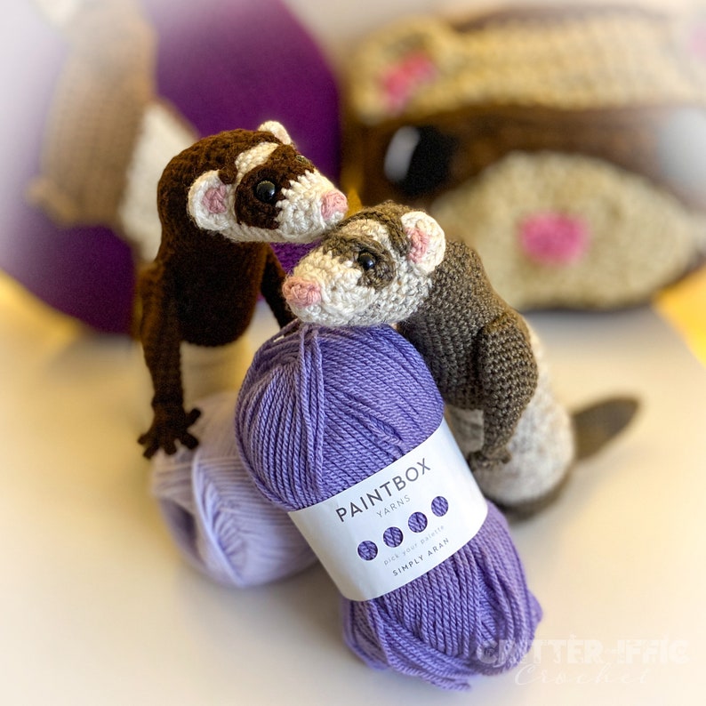 two crocheted ferrets propped up on purple yarn