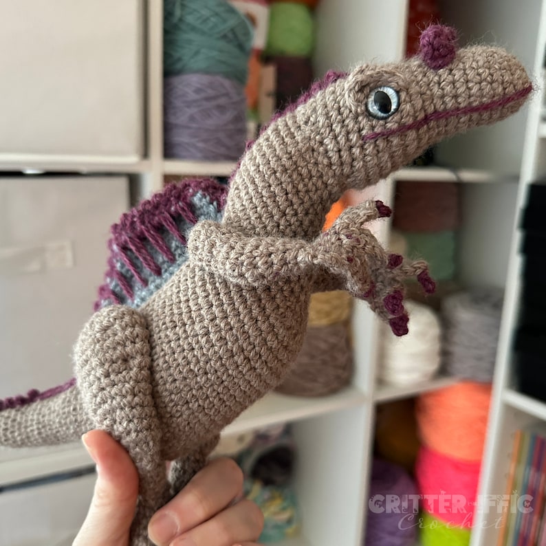Pode incluir: Um brinquedo de dinossauro de croch&ecirc; feito &agrave; m&atilde;o em tons de bege, roxo e azul. O dinossauro tem um acabamento texturizado, com garras detalhadas e uma barbatana distinta nas costas. Os olhos s&atilde;o grandes e redondos. O texto "Critterrific Crochet" &eacute; vis&iacute;vel.