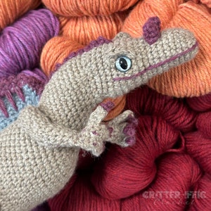 Pode incluir: Um brinquedo de dinossauro de croch&ecirc; em tons de bege, roxo e azul, com um design detalhado. O dinossauro est&aacute; sobre um fundo de fios coloridos em tons de laranja, roxo e vermelho. O brinquedo tem uma express&atilde;o amig&aacute;vel.