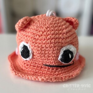 Puede incluir: Un sombrero de cubo de punto naranja con ojos negros, detalles blancos en los ojos y una boca cosida negra. El sombrero tiene una cuerda de punto blanca en la parte superior.