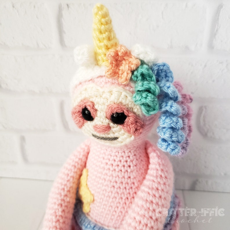 close up of crocheted sloth in unicorn costume face against white brick background