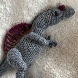 Pode incluir: Um brinquedo de dinossauro de croch&ecirc; em tons de cinza, com uma estrutura em forma de vela bord&ocirc; e bege nas costas. O dinossauro tem olhos pequenos e escuros e uma crista bord&ocirc;. O brinquedo &eacute; feito de fio macio e est&aacute; sobre uma superf&iacute;cie branca e fofa.