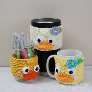 May include: Three crocheted duck-shaped cup cozies in yellow, white, and light yellow. The cozies have black eyes, orange beaks, and pink cheeks. The cozies are on a white surface.