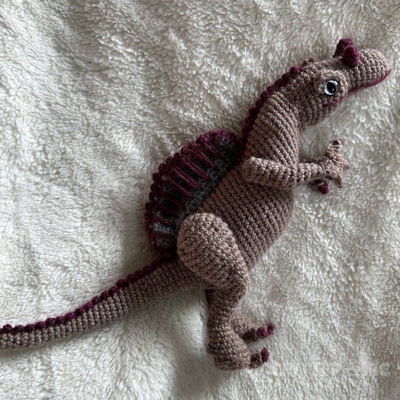 Pode incluir: Um brinquedo de dinossauro de croch&ecirc; em tons de marrom, com uma espinha dorsal e detalhes em bord&ocirc; e cinza. O dinossauro tem p&eacute;s pequenos e detalhados e uma cauda longa. Os olhos s&atilde;o pretos e o brinquedo &eacute; feito de fio macio.