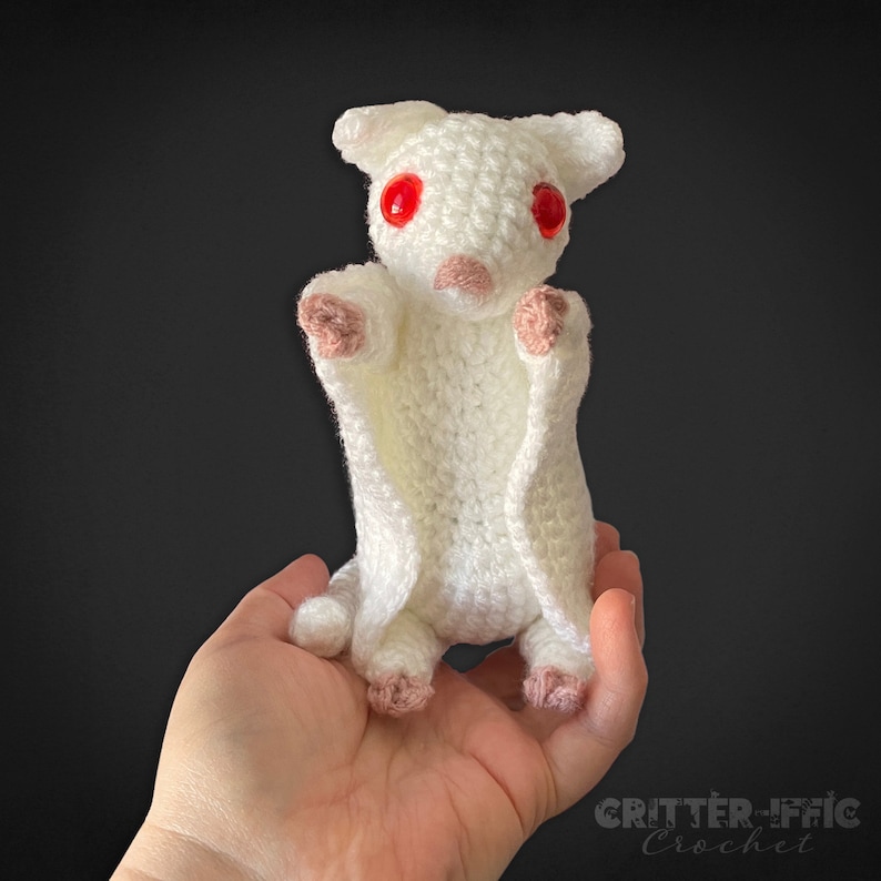 white crocheted sugar glider with red eyes being held by a hand on a black background