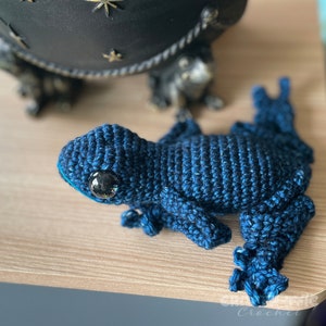 crocheted red eyed tree frog made in blue with black eyes sitting on a table next to a cauldron