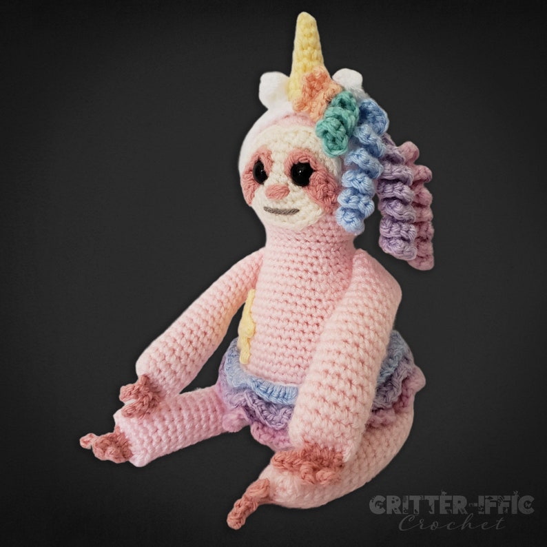front left side view of crocheted pink sloth in unicorn costume on a black background