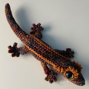 a curious brown crocheted crested gecko sitting on a white table looking up at the camera
