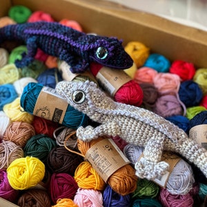 2 crocheted crested geckos, one purple and one smokey grey sitting in a box of yarn