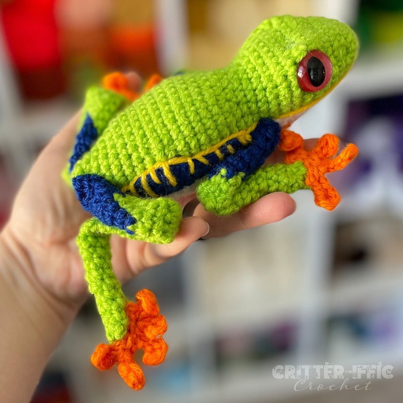 May include: A crocheted green frog with orange feet and blue and yellow details. The frog is being held by a hand.