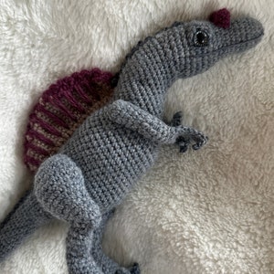 Pode incluir: Um dinossauro de croch&ecirc; feito &agrave; m&atilde;o em tons de cinza, com uma vela bord&ocirc; e bege nas costas. O dinossauro tem pequenos membros detalhados e uma crista bord&ocirc;. O brinquedo est&aacute; sobre uma superf&iacute;cie macia e branca.