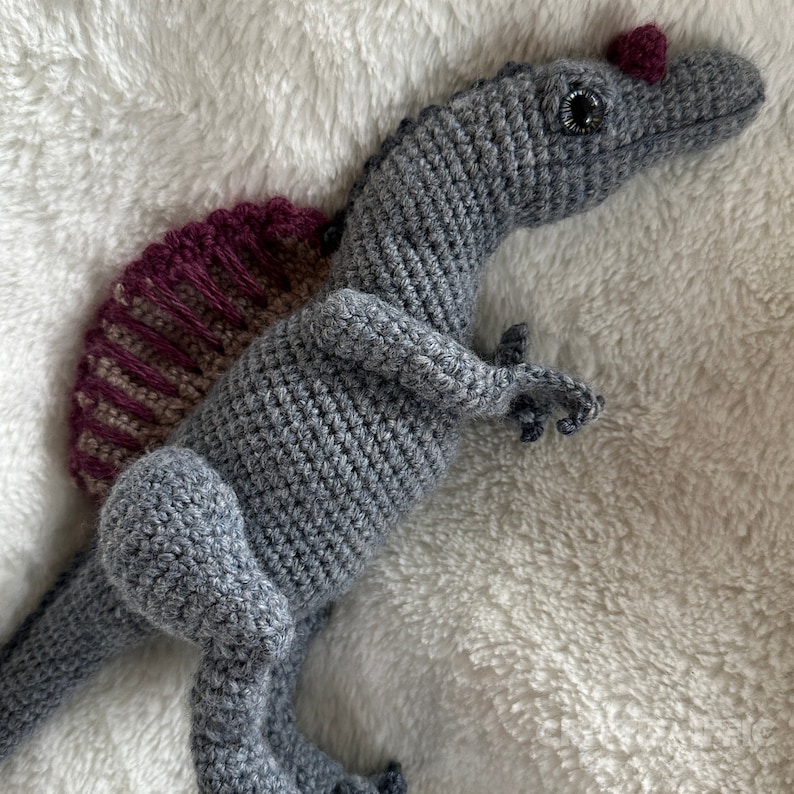 Pode incluir: Um dinossauro de croch&ecirc; feito &agrave; m&atilde;o em tons de cinza, com uma vela bord&ocirc; e bege nas costas. O dinossauro tem pequenos membros detalhados e uma crista bord&ocirc;. O brinquedo est&aacute; sobre uma superf&iacute;cie macia e branca.