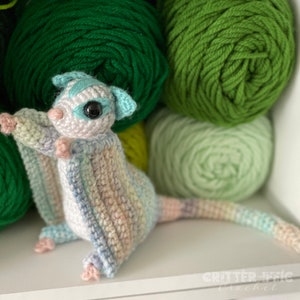 rainbow multicolored crocheted sugar gldier sitting on a white shelf in front of green yarn