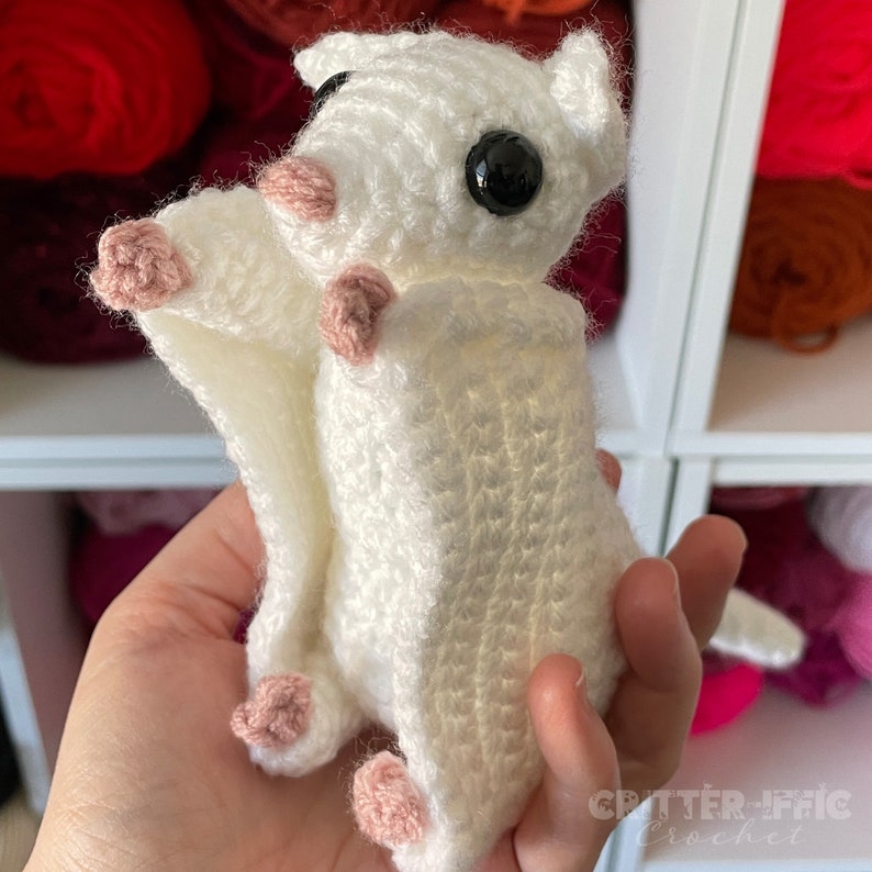 white crocheted sugar glider being held in front of a shelf full of red yarn