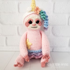 crocheted sloth in a unicorn costume against a white brick background facing front