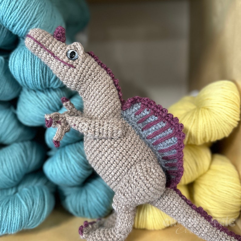 Pode incluir: Um brinquedo de dinossauro Spinosaurus de croch&ecirc; em tons de bege, cinza e roxo. O dinossauro feito &agrave; m&atilde;o tem uma vela distinta nas costas e posa com os bra&ccedil;os levantados. O fundo inclui novelos de l&atilde; turquesa e amarela.