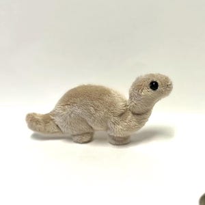 May include: A soft, beige turtle toy with a detailed, textured finish. The turtle has a small head, a curved neck, and a short tail. Small, smooth stones are in the background. The toy is a decorative item.