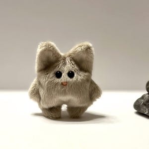 May include: A small, grey, plush toy with large black eyes and a pink nose. The toy is shaped like a cat and has a fluffy texture.