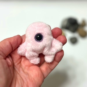 Peut inclure: Un petit jouet en peluche rose avec un seul gros œil noir. Le jouet a quatre petites pattes et un corps rond.