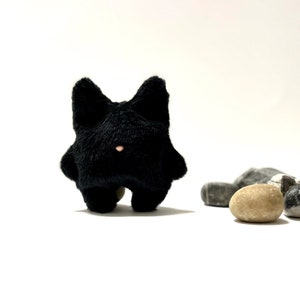 May include: A black plush toy cat with a pink nose, standing on a white surface with grey and tan stones.