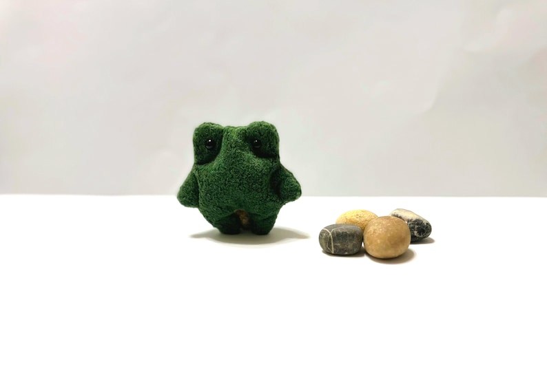 K&ouml;nnte beinhalten: Ein kleiner gr&uuml;ner Pl&uuml;schfrosch mit schwarzen Augen sitzt auf einer wei&szlig;en Oberfl&auml;che neben vier glatten Steinen. Zwei der Steine sind hellbraun und zwei sind dunkelgrau.