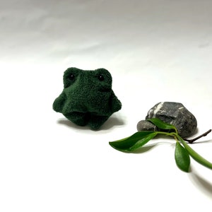 K&ouml;nnte beinhalten: Ein kleiner, gr&uuml;ner Pl&uuml;schfrosch sitzt auf einer wei&szlig;en Oberfl&auml;che. Ein kleiner, grauer Stein mit wei&szlig;en Streifen befindet sich rechts vom Frosch. Ein kleines gr&uuml;nes Blatt liegt vor dem Stein.