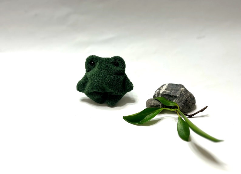 K&ouml;nnte beinhalten: Ein kleiner, gr&uuml;ner Pl&uuml;schfrosch sitzt auf einer wei&szlig;en Oberfl&auml;che. Ein kleiner, grauer Stein mit wei&szlig;en Streifen befindet sich rechts vom Frosch. Ein kleines gr&uuml;nes Blatt liegt vor dem Stein.